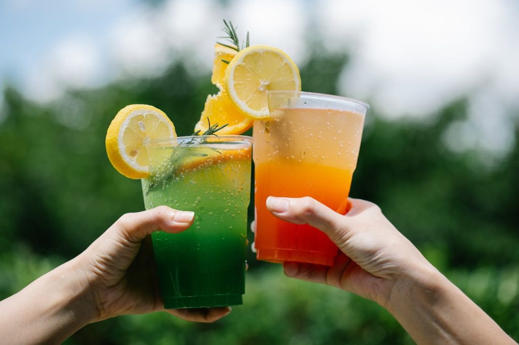 Two hands holding colorful citrus lemonades in a vibrant summer outdoor setting.